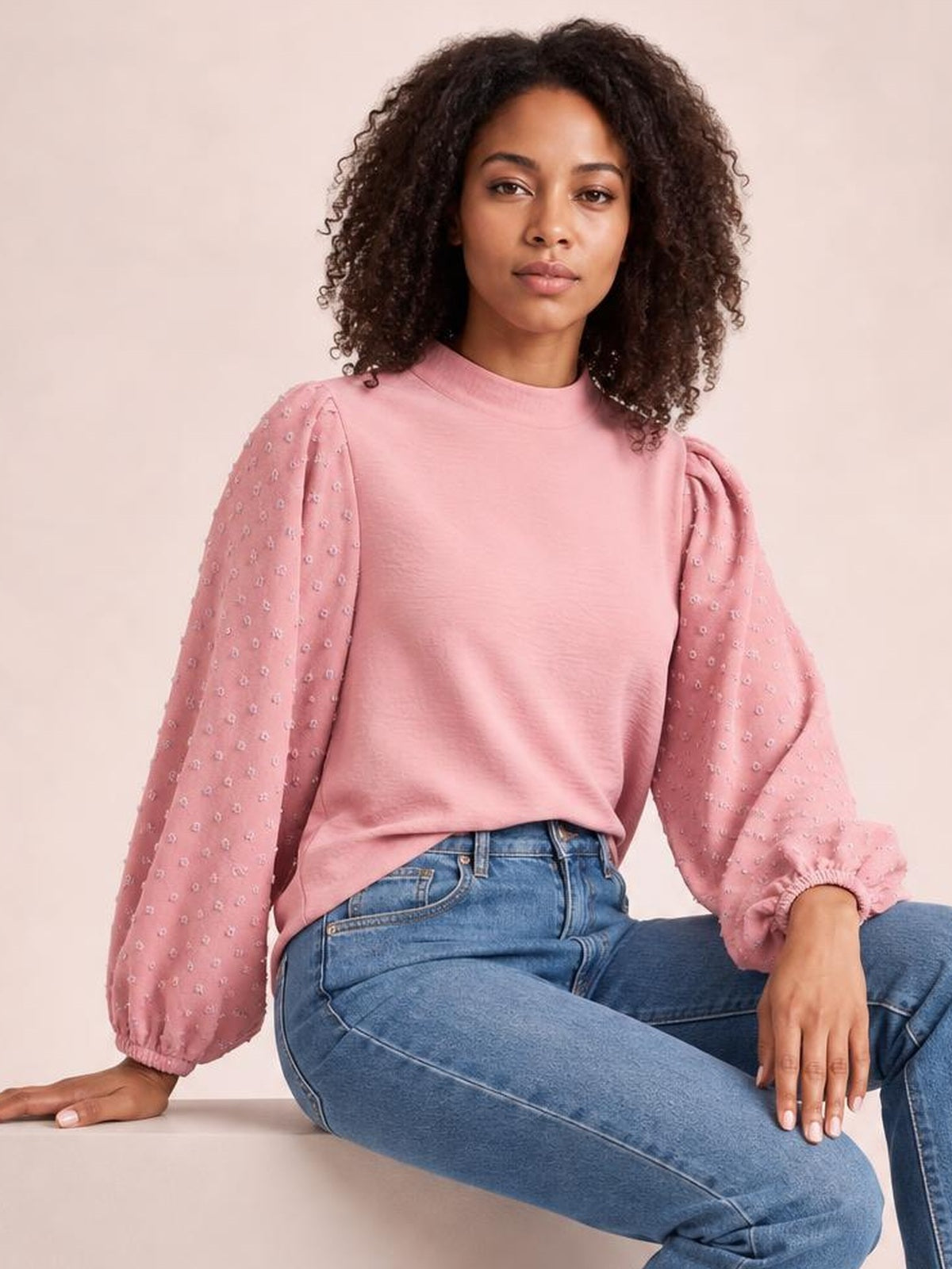 Model seated in a pink Swiss dot mock neck blouse with voluminous balloon sleeves, gathered cuffs, and a relaxed silhouette.
