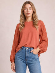 A model wears a rust Swiss dot mock neck blouse with voluminous balloon sleeves, gathered cuffs, and a relaxed silhouette, posing with one hand in her jeans pocket.