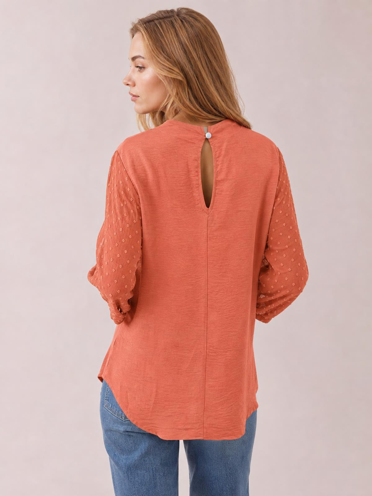 A model shows the back of a coral Swiss dot mock neck blouse with balloon sleeves, a buttoned keyhole opening, curved hem, and relaxed silhouette.