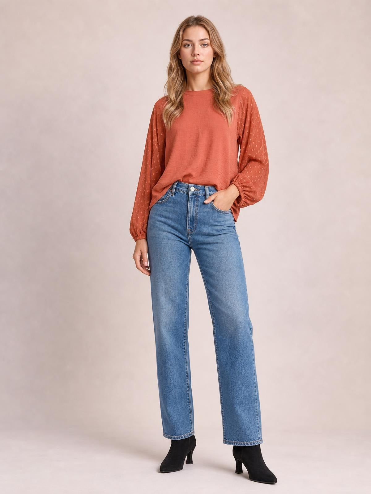 Model wears a rust Swiss dot mock neck blouse with voluminous balloon sleeves and gathered cuffs, styled in a relaxed silhouette with one hand in her jeans pocket.