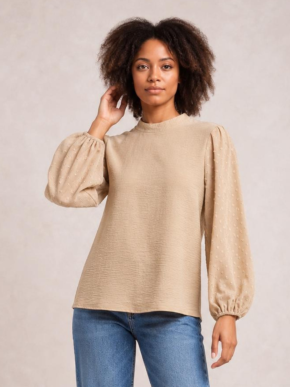 A model poses in a beige Swiss dot mock neck blouse with a relaxed straight silhouette, voluminous balloon sleeves, gathered cuffs, and a softly textured finish.