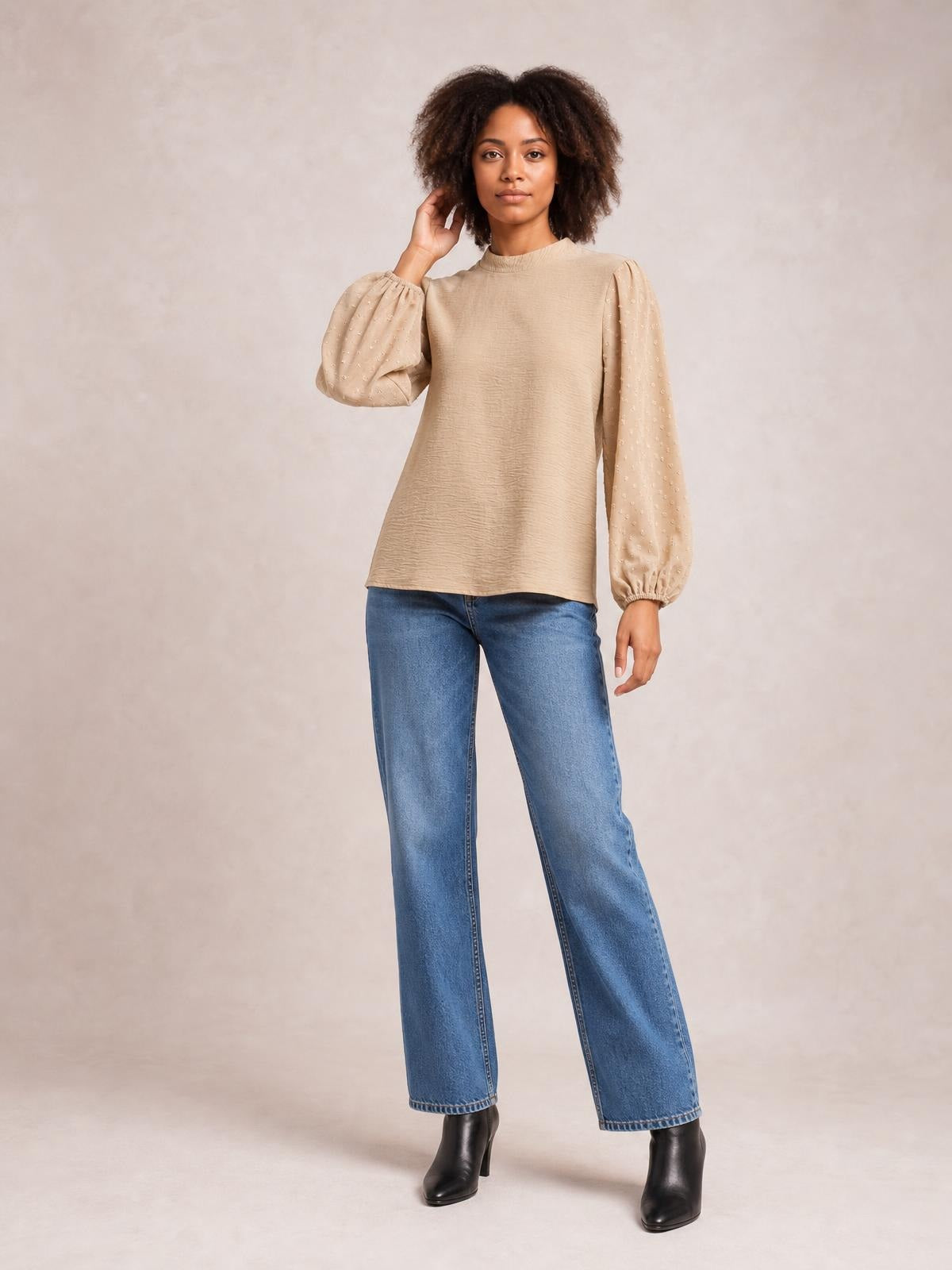 Model wearing a beige Swiss dot mock neck blouse with voluminous balloon sleeves, gathered cuffs, and a relaxed straight silhouette, posing with one hand in her hair.