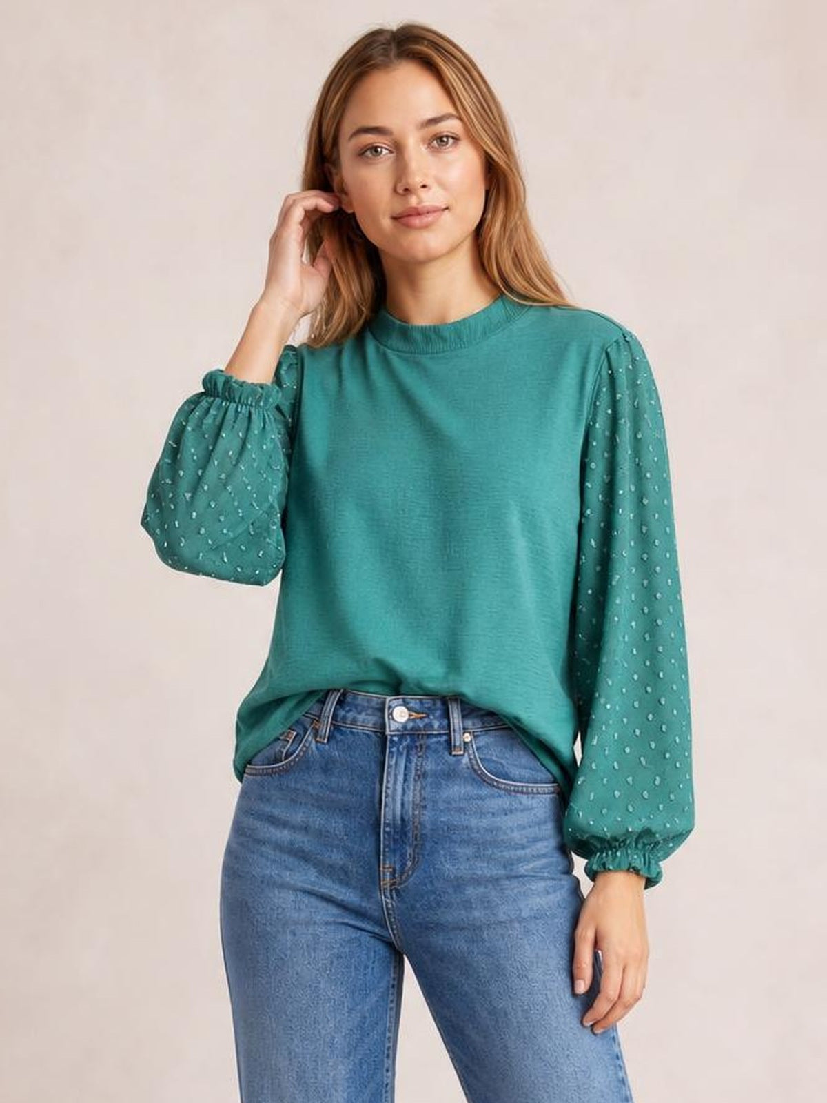 Model wearing a teal Swiss dot mock neck blouse with voluminous balloon sleeves, gathered cuffs, and a relaxed silhouette, touching her hair.