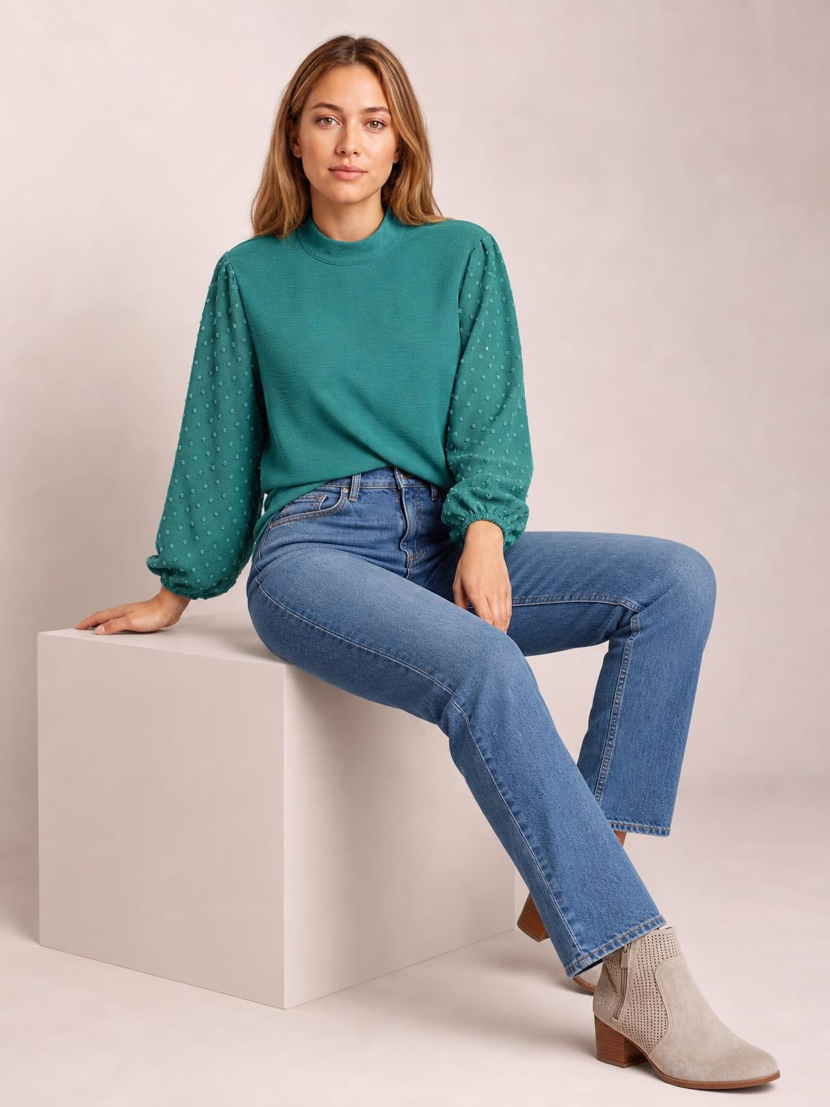 Model seated in a teal Swiss dot mock neck blouse with voluminous balloon sleeves, gathered cuffs, and a relaxed silhouette.