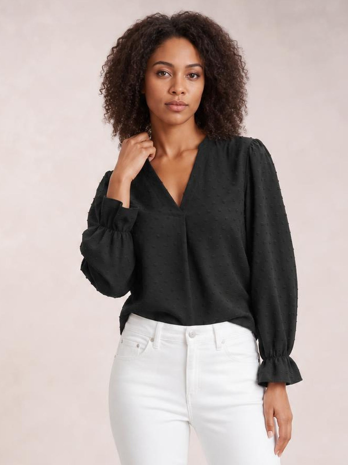 Model wearing a black Swiss dot blouse with a notched V-neck, relaxed silhouette, long flounce sleeves, gathered cuffs, and a gently curved hem.