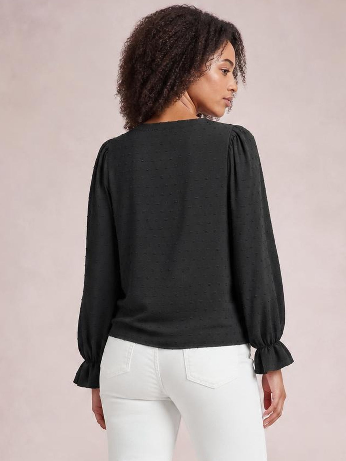 A model looks over her shoulder wearing a black Swiss dot blouse with a relaxed long silhouette, long sleeves, gathered cuffs, and flounce detailing.