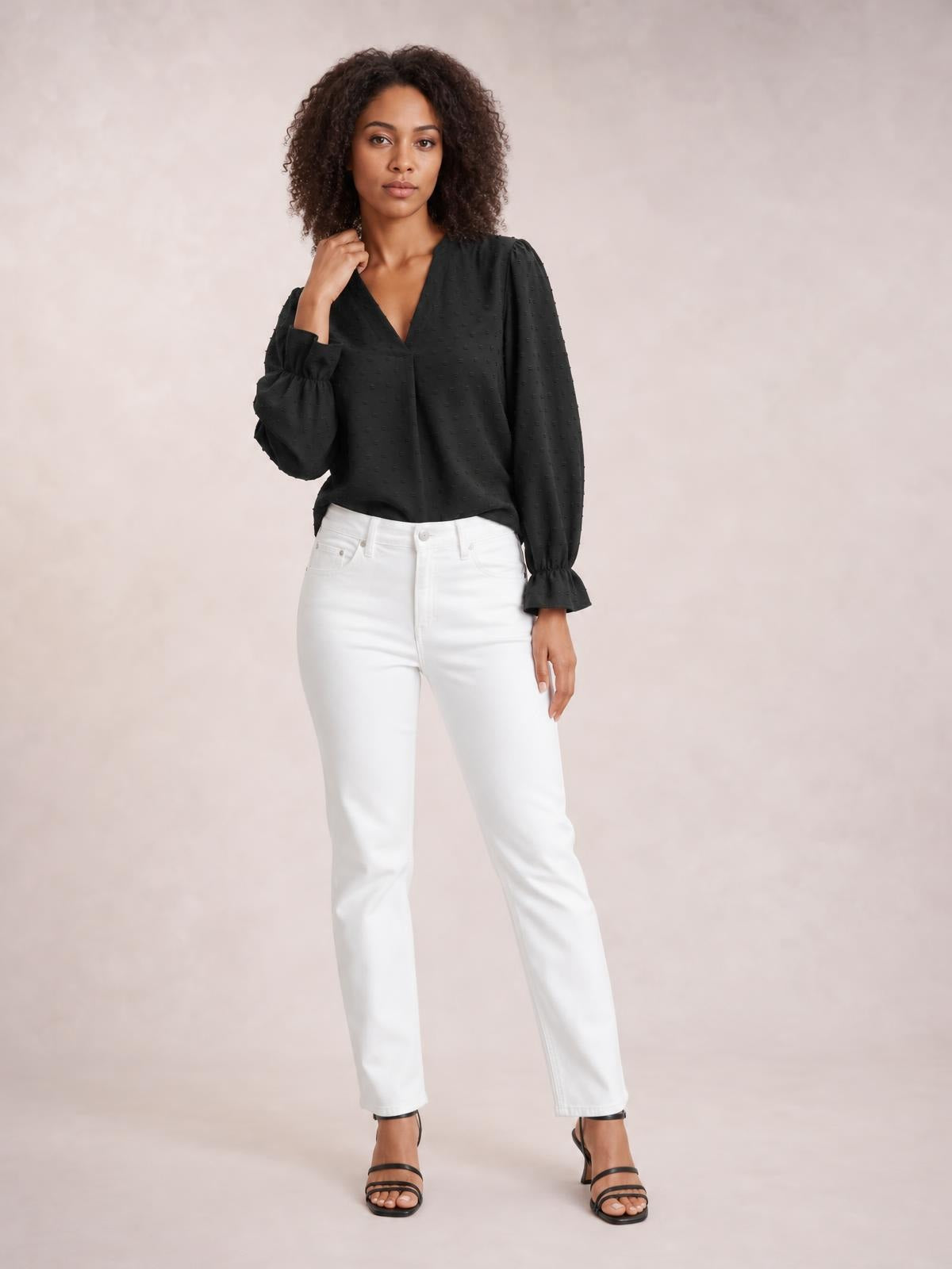 A model poses in a black Swiss dot blouse with a notched V-neck, long flounce sleeves, gathered cuffs, and a relaxed long silhouette.