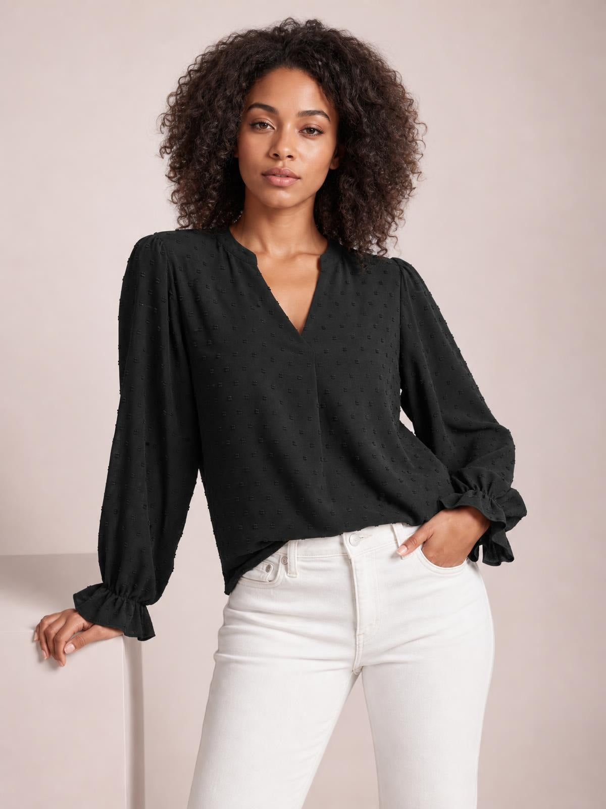 A model poses in a black Swiss dot blouse with a notched V-neck, relaxed silhouette, long sleeves, and flounce cuffs, paired with white jeans.