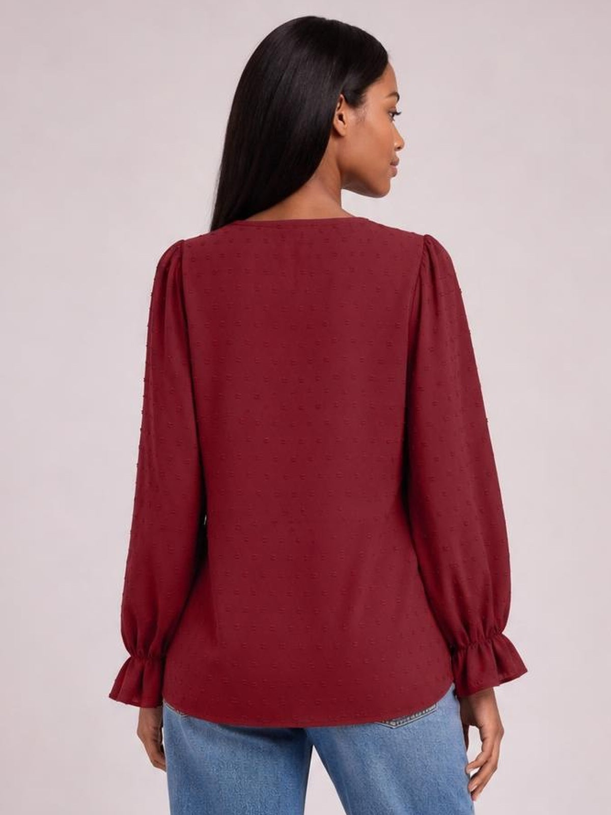 A model shows the back of a burgundy Swiss dot blouse with a relaxed long silhouette, long sleeves, and flounce cuffs.