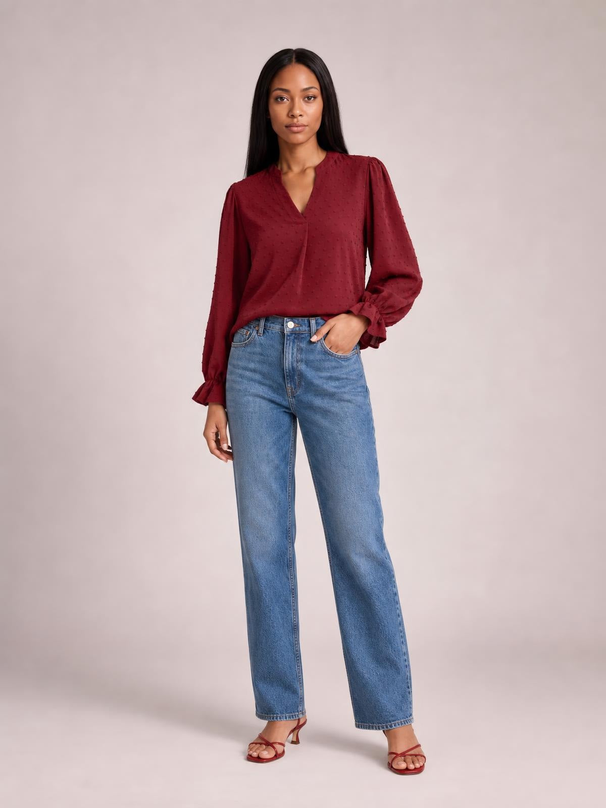 Model poses with one hand in her jeans pocket wearing a burgundy Swiss dot blouse with a notched V-neck, long flounce sleeves, and a relaxed silhouette.