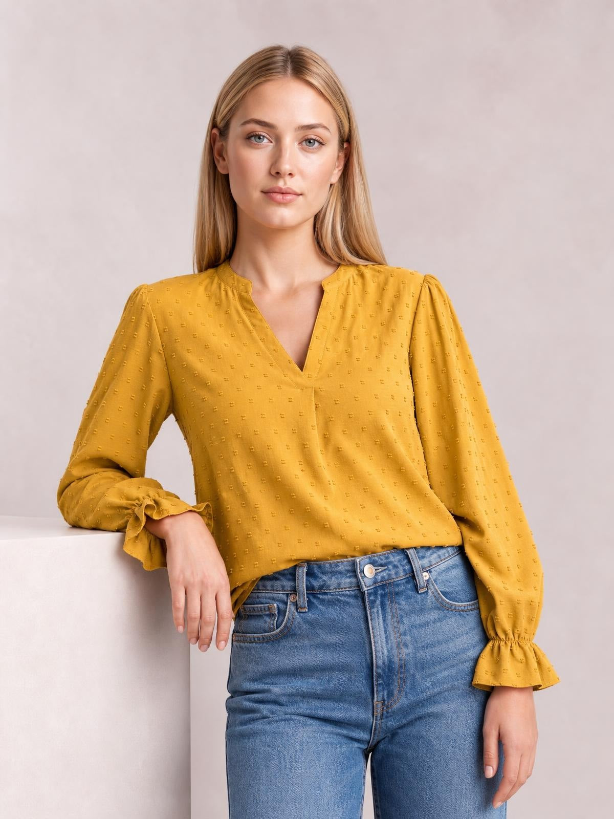 A model wears a mustard Swiss dot blouse with a notched V-neck, relaxed silhouette, long sleeves, and flounced cuffs while leaning with one hand.