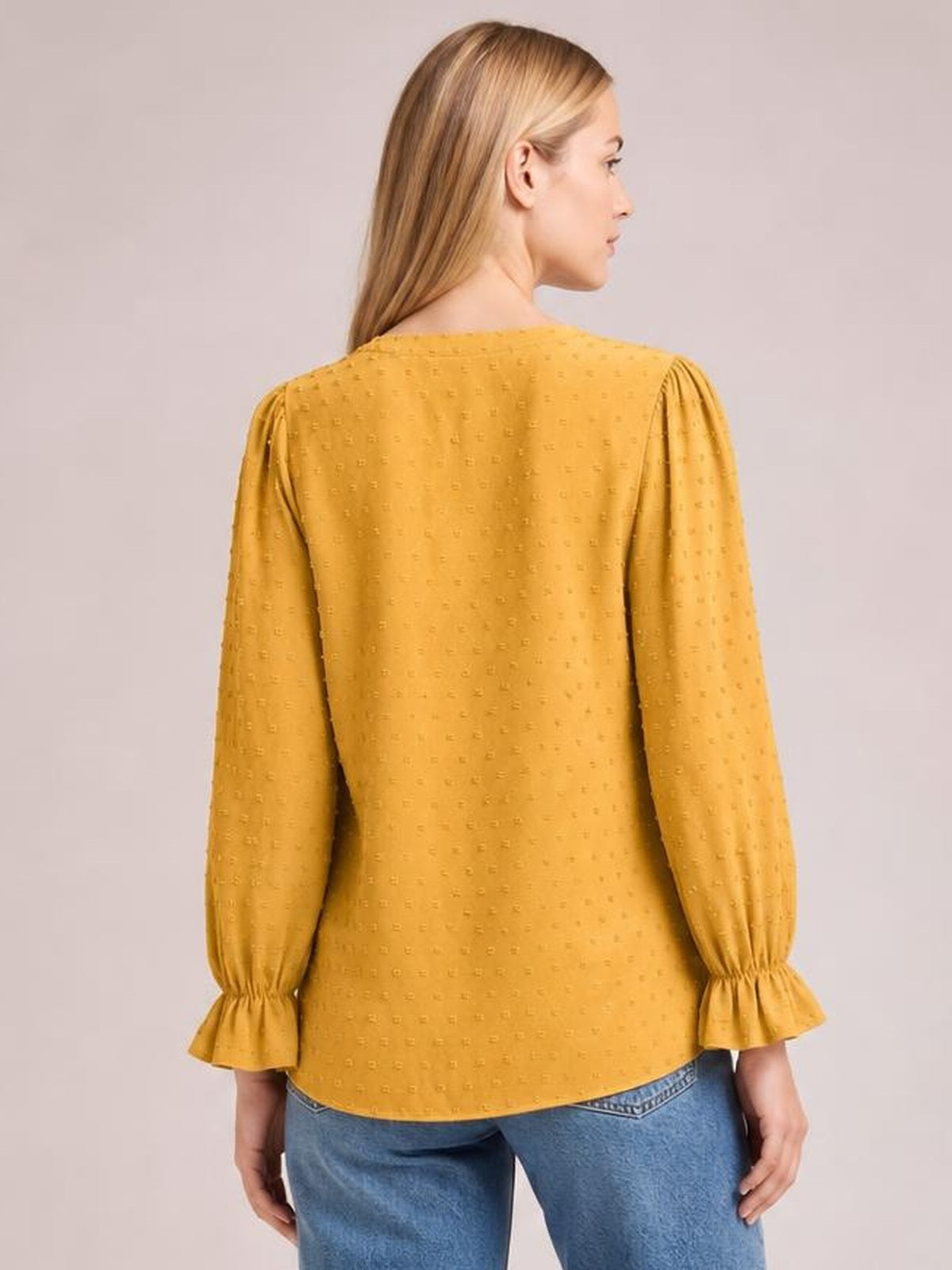 A model shows the back of a mustard yellow Swiss dot blouse with a relaxed long silhouette, full sleeves, gathered cuffs, and flounced hem details.