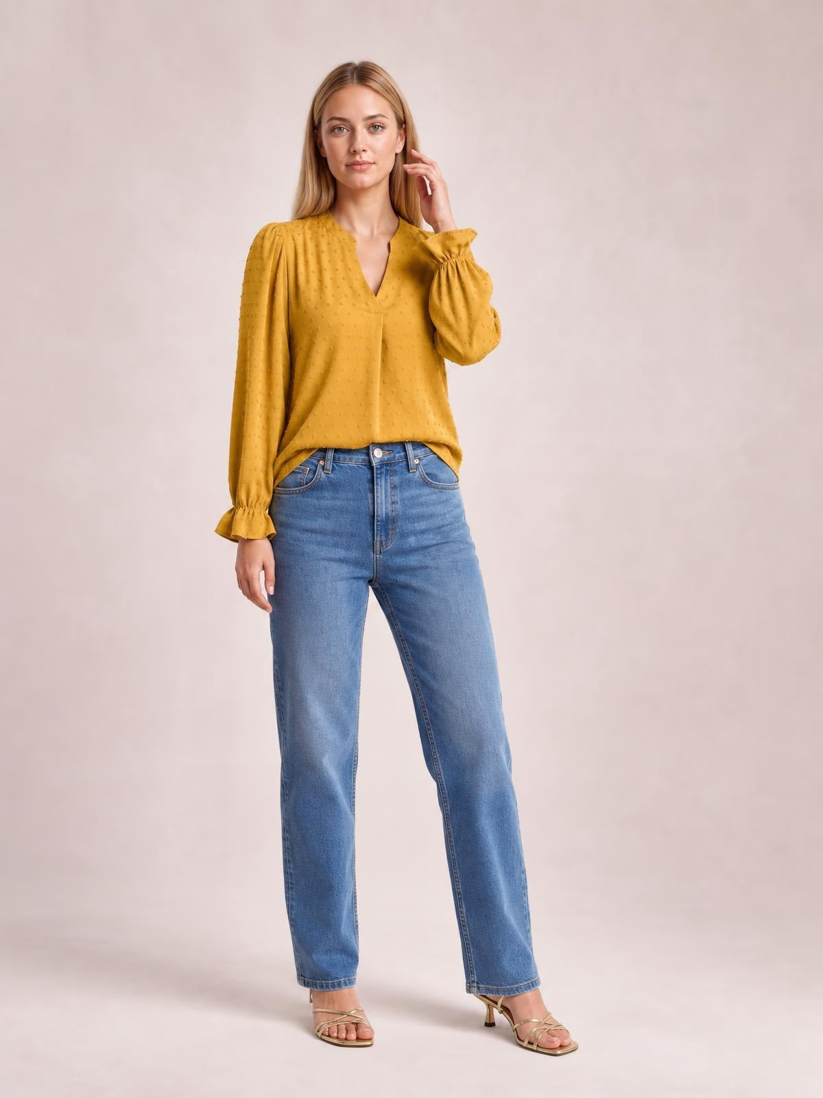Model wearing a mustard Swiss dot blouse with a notched V-neck, long flounce sleeves, gathered cuffs, and a relaxed long silhouette with jeans.