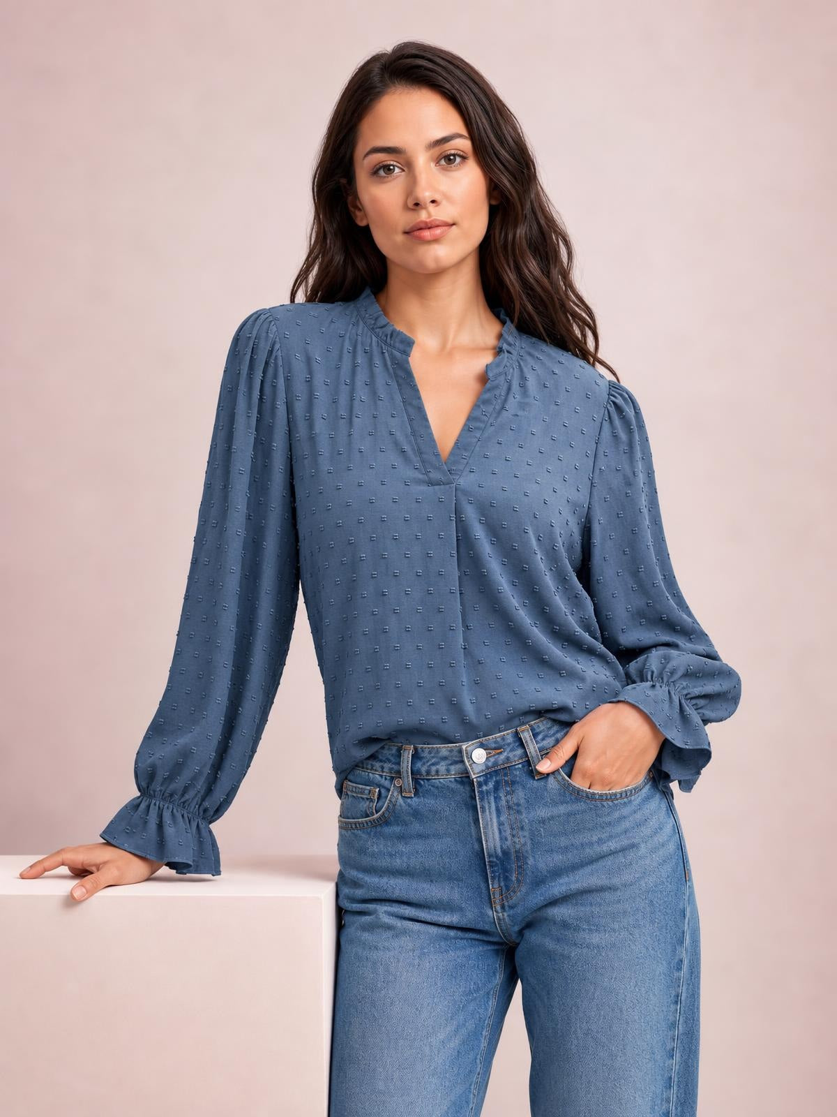 A model wears a solid blue Swiss dot blouse with a notched V-neck, relaxed long silhouette, and gathered flounce sleeves, posing with one hand in her jeans pocket.