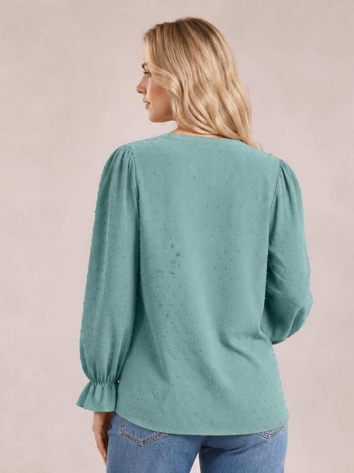 A model turns slightly away in a relaxed teal Swiss dot blouse with long flounce sleeves, gathered shoulders, and a gently curved hem.