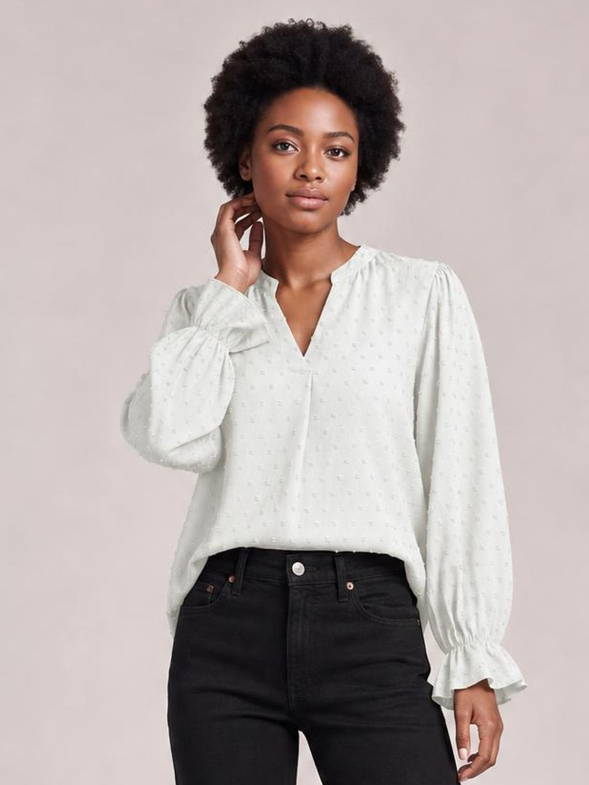 A model wears a white Swiss dot blouse with a notched V-neck, long flounce sleeves, gathered cuffs, and a relaxed long silhouette while touching her neck.