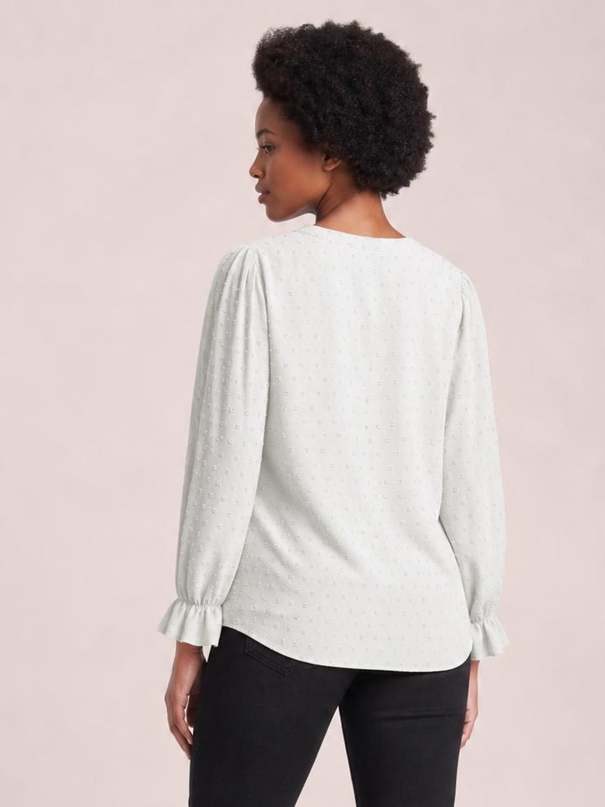 A model poses turned away in a white Swiss dot blouse with a relaxed long silhouette, long sleeves, and gathered flounce cuffs.