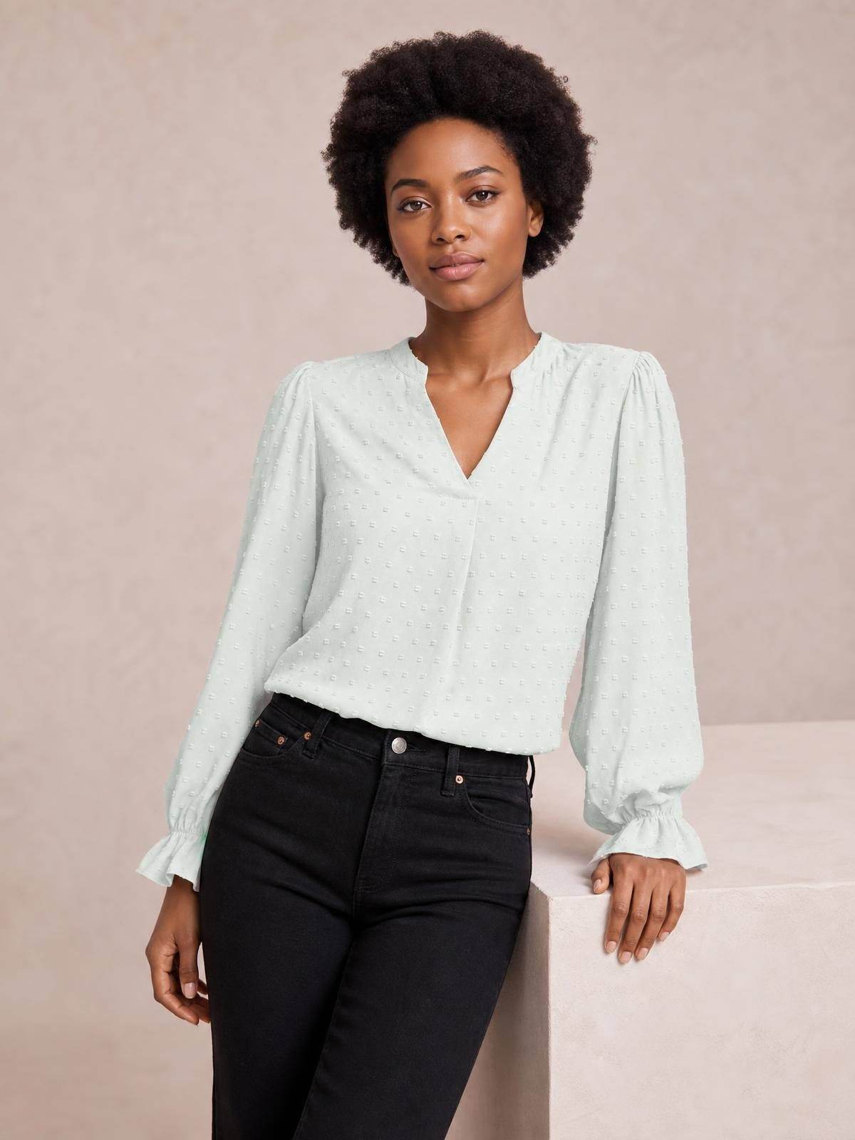 A model leans against a block wearing a pale mint Swiss dot blouse with a notched V-neck, relaxed silhouette, and long flounce sleeves.