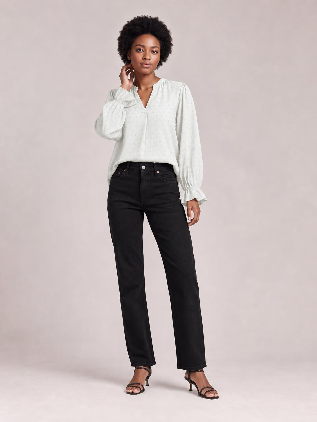 A model touches her neck while wearing a white Swiss dot blouse with a notched V-neck, relaxed long-sleeve silhouette, and gathered flounce cuffs.