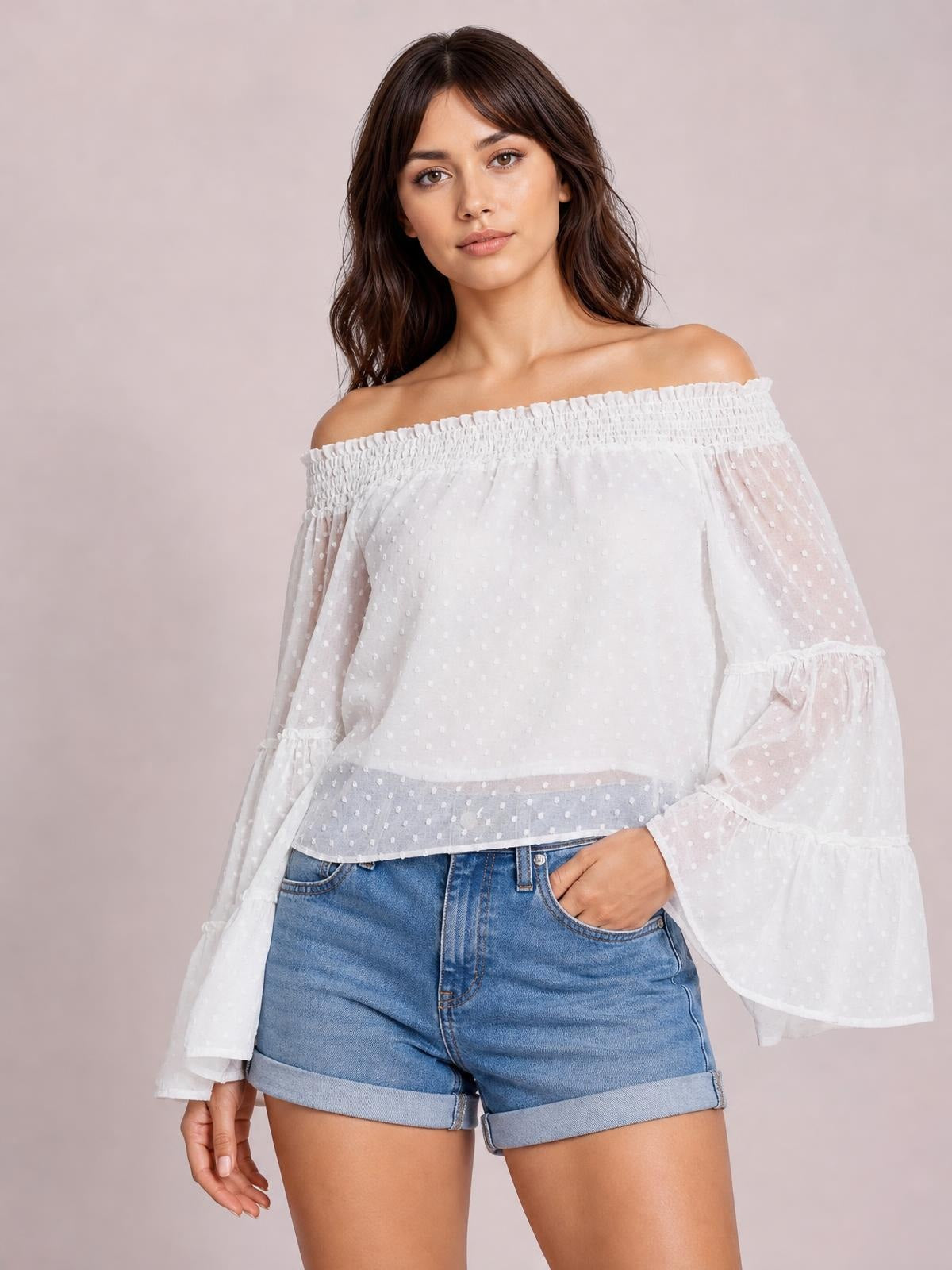 Model poses with one hand in her pocket wearing a white Swiss dot off-shoulder blouse with a smocked neckline, sheer flared long sleeves, and frilled cuffs.