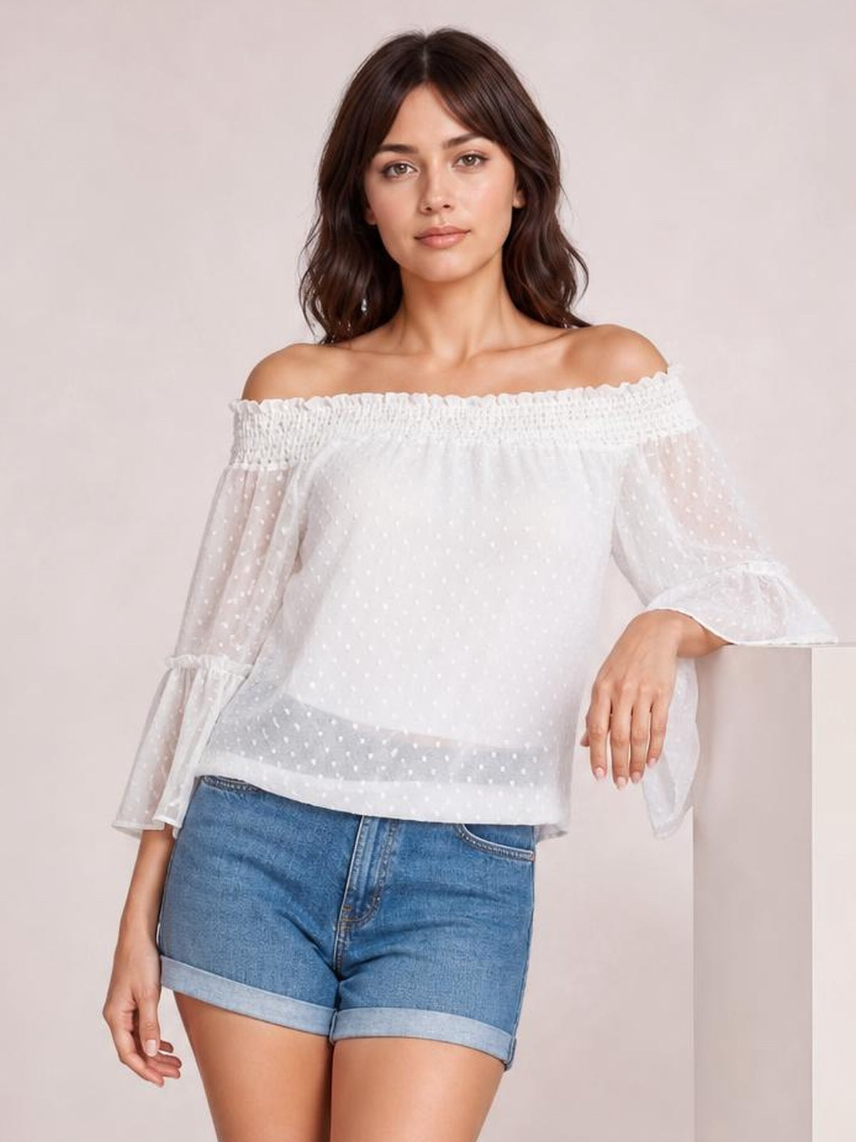 A model wears a white Swiss dot off-shoulder blouse with a frilled neckline, sheer lined body, long flare sleeves, and a relaxed regular-length silhouette.