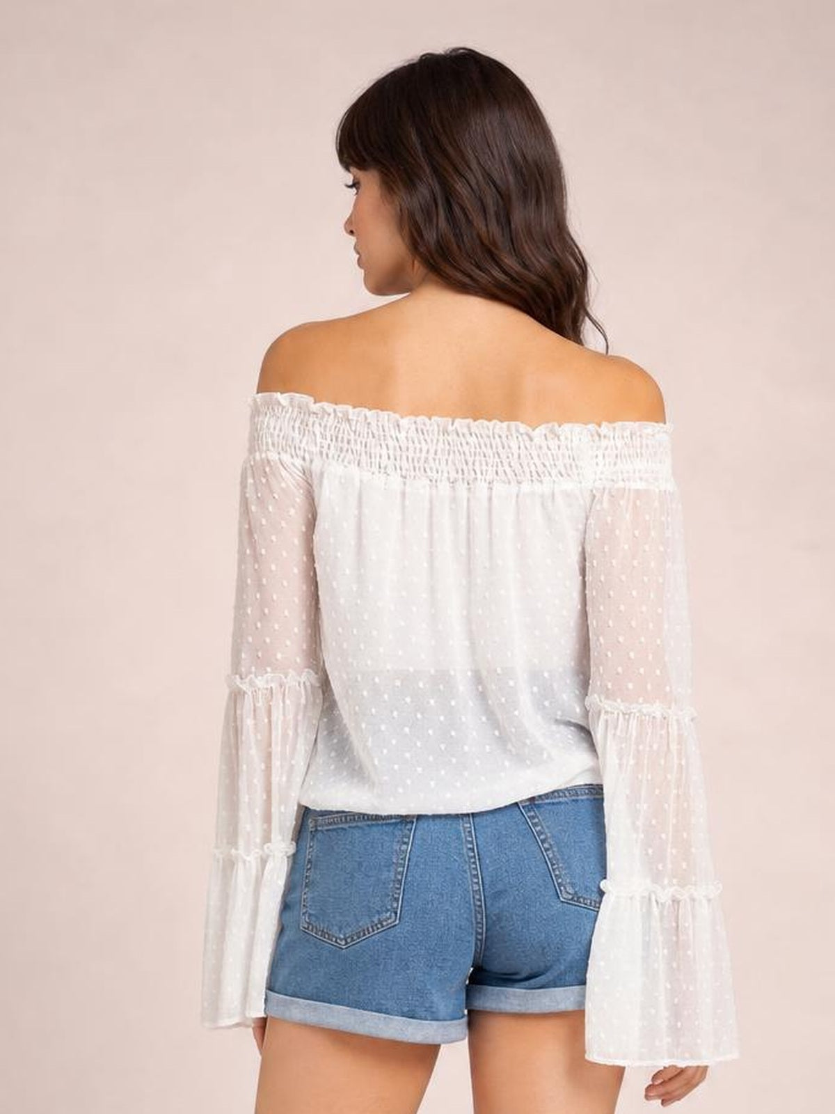 Model shown from behind wears a white Swiss dot off-shoulder blouse with an elasticized neckline, relaxed silhouette, and long sheer flare sleeves with frilled cuffs.