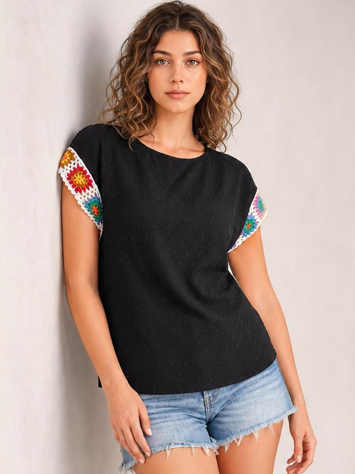 A model wears a black Swiss dot top with a round neckline, short sleeves trimmed in colorful crochet, and a relaxed, straight silhouette.