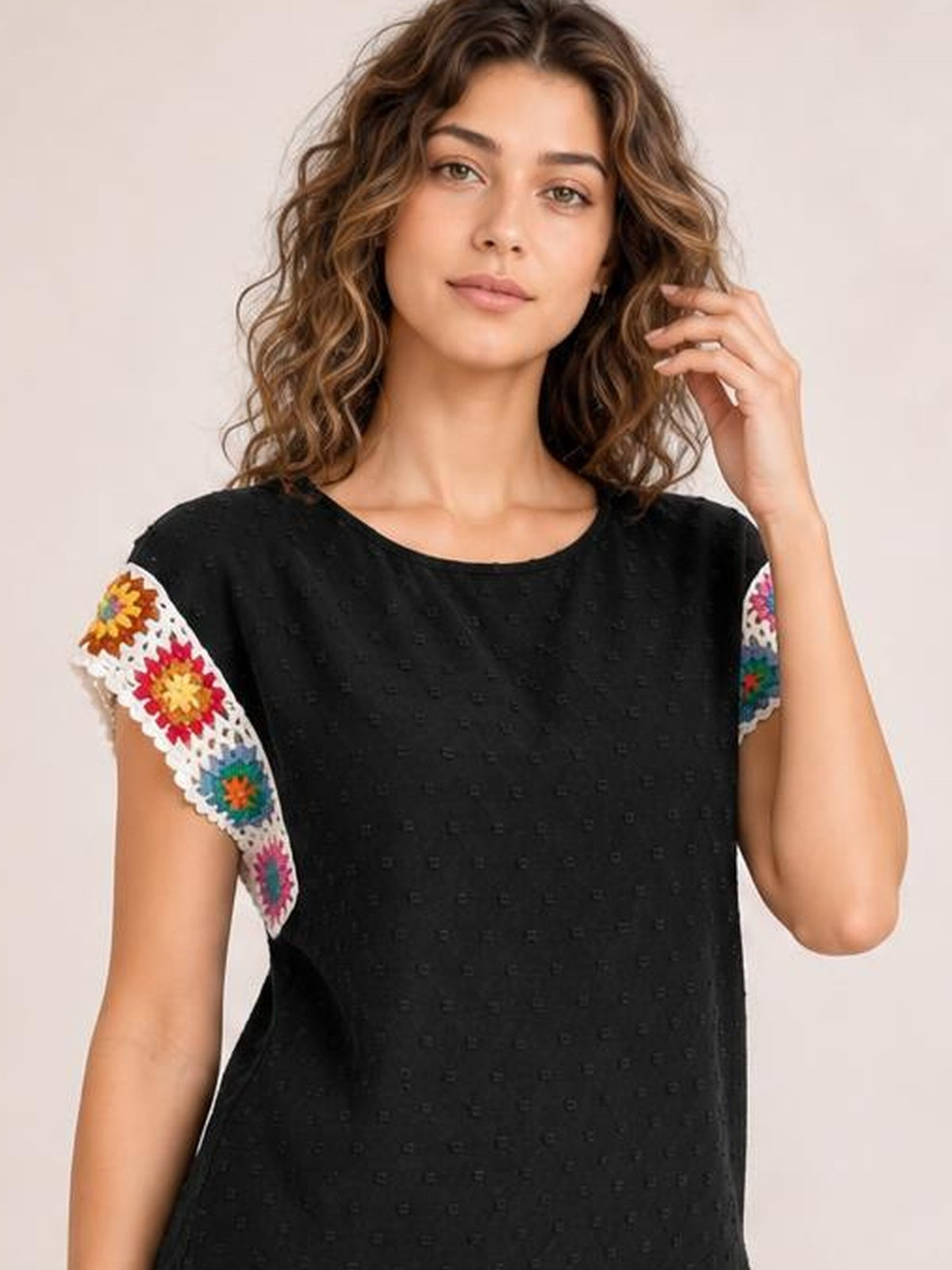 Model poses with one hand in her hair, wearing a black Swiss dot round-neck top with a relaxed silhouette and colorful crochet floral short sleeves.