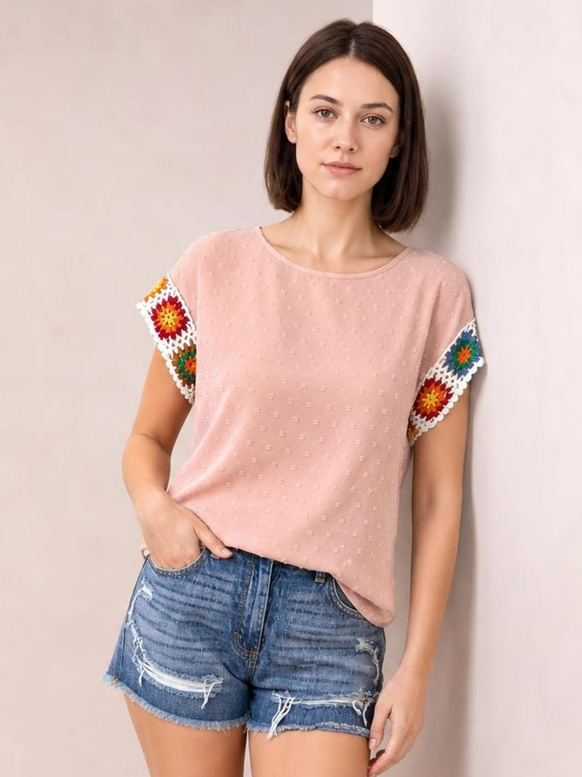 Model wears a blush pink Swiss dot round-neck top with a relaxed silhouette, short sleeves, and colorful embroidered crochet trim, posing with one hand in her pocket.