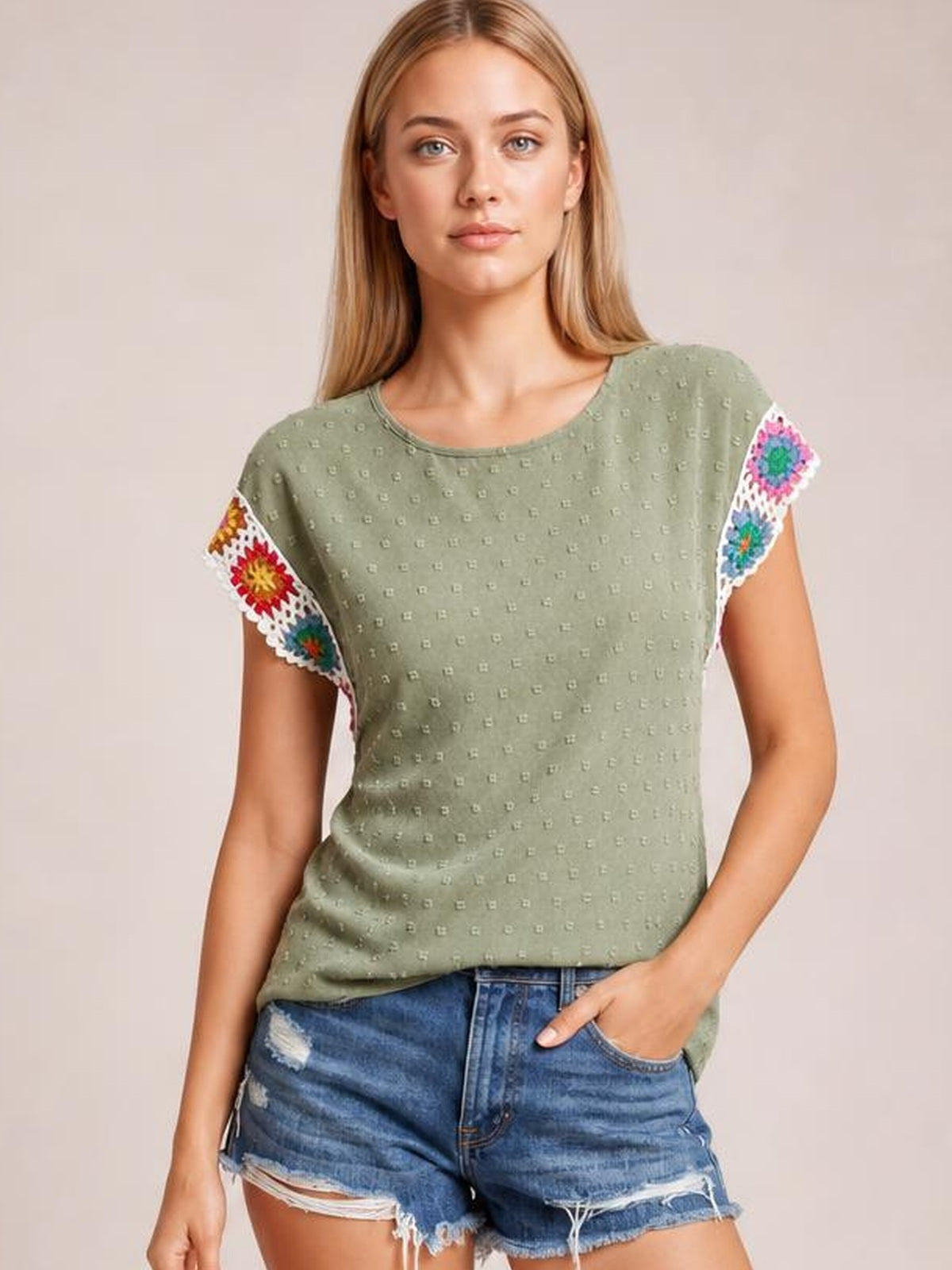 A model wears an olive green Swiss dot top with a round neck, short sleeves trimmed in colorful crochet floral panels, and a relaxed straight silhouette.