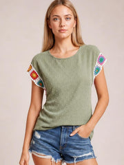 A model wears an olive green Swiss dot top with a round neck, short sleeves trimmed in colorful crochet floral panels, and a relaxed straight silhouette.