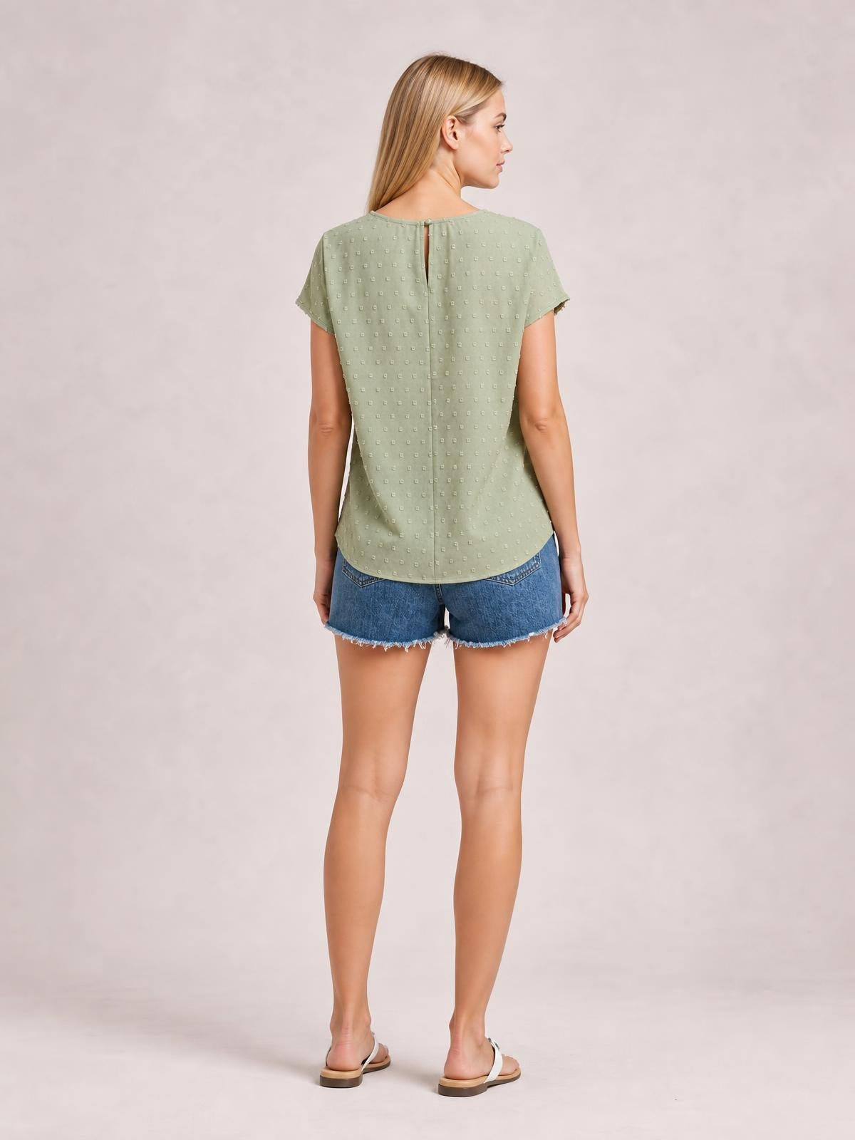 Model shown from the back in a sage green Swiss dot crochet top with short sleeves, a round neck, buttoned back opening, and curved hem.