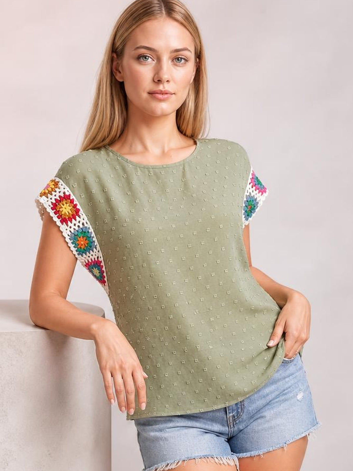 A model leans casually in a sage green Swiss dot top with a round neck, short sleeves, colorful crochet floral trim, and a relaxed straight silhouette.