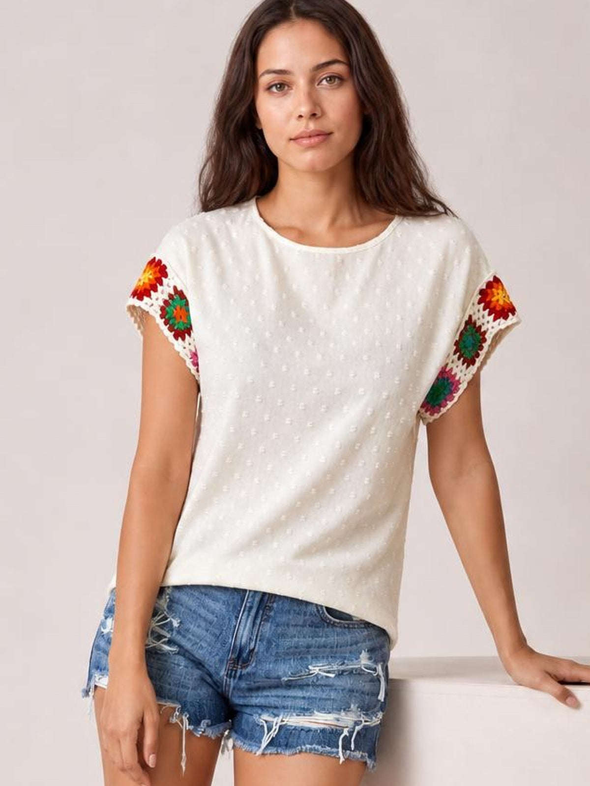 A model wears an ivory Swiss dot top with a round neck, relaxed silhouette, short sleeves, and colorful embroidered crochet trim at the cuffs.
