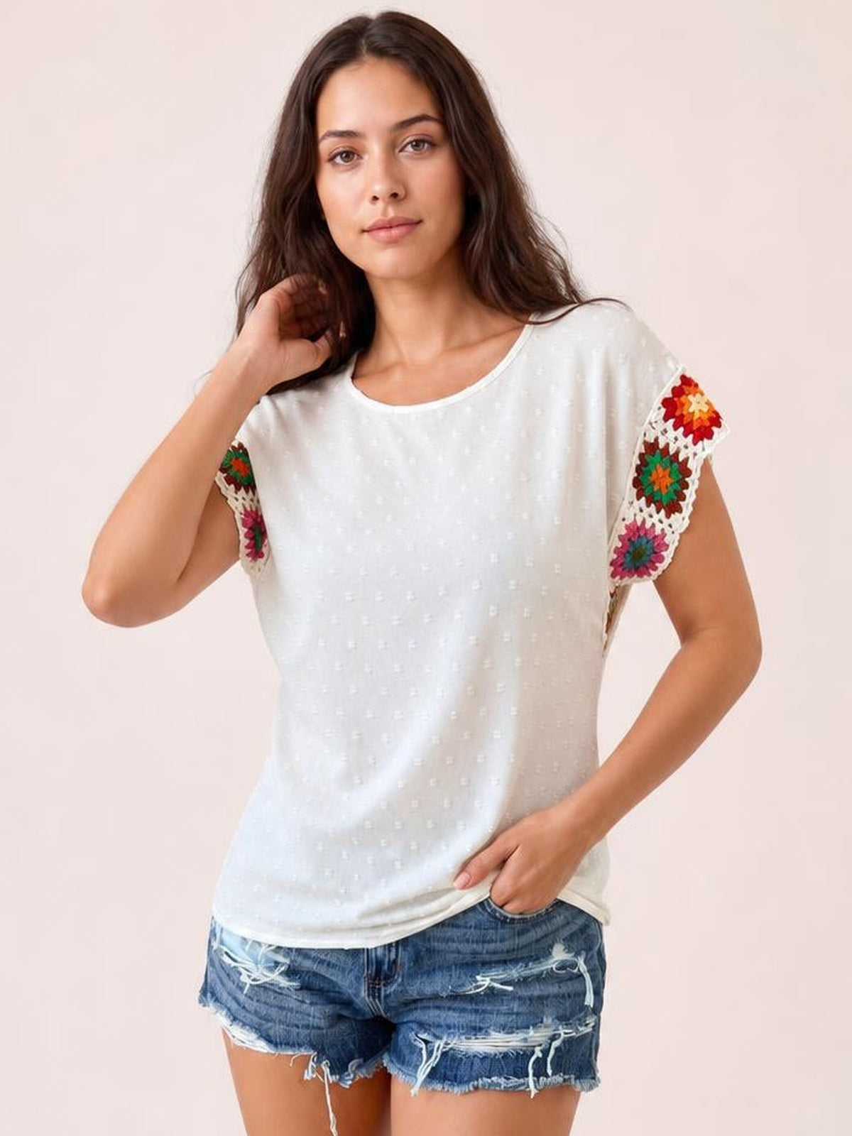 A woman poses in a white Swiss dot top with a round neck, short sleeves, colorful crochet floral sleeve panels, and a relaxed straight silhouette.