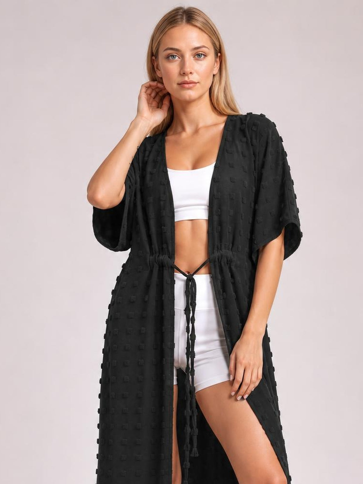 A model poses in a black Swiss dot sheer beach cover-up maxi dress with wide sleeves, an open front, tied waist, and high split over white swimwear.