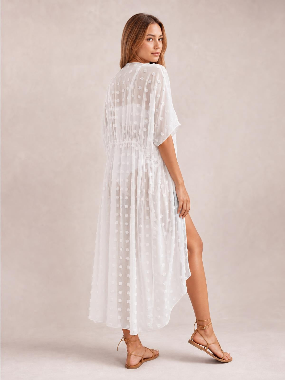 Model walks in a white sheer Swiss dot beach cover-up maxi dress with short sleeves, tied waist, flowing silhouette, and high side split.