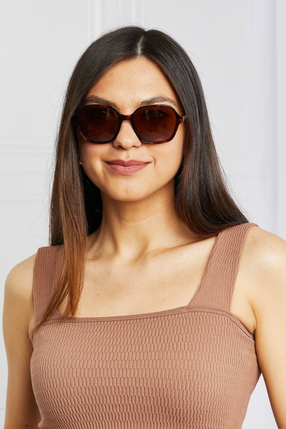 Model faces forward wearing brown full-rim square sunglasses with softly rounded edges, dark warm-toned lenses, and a sculpted acetate frame.