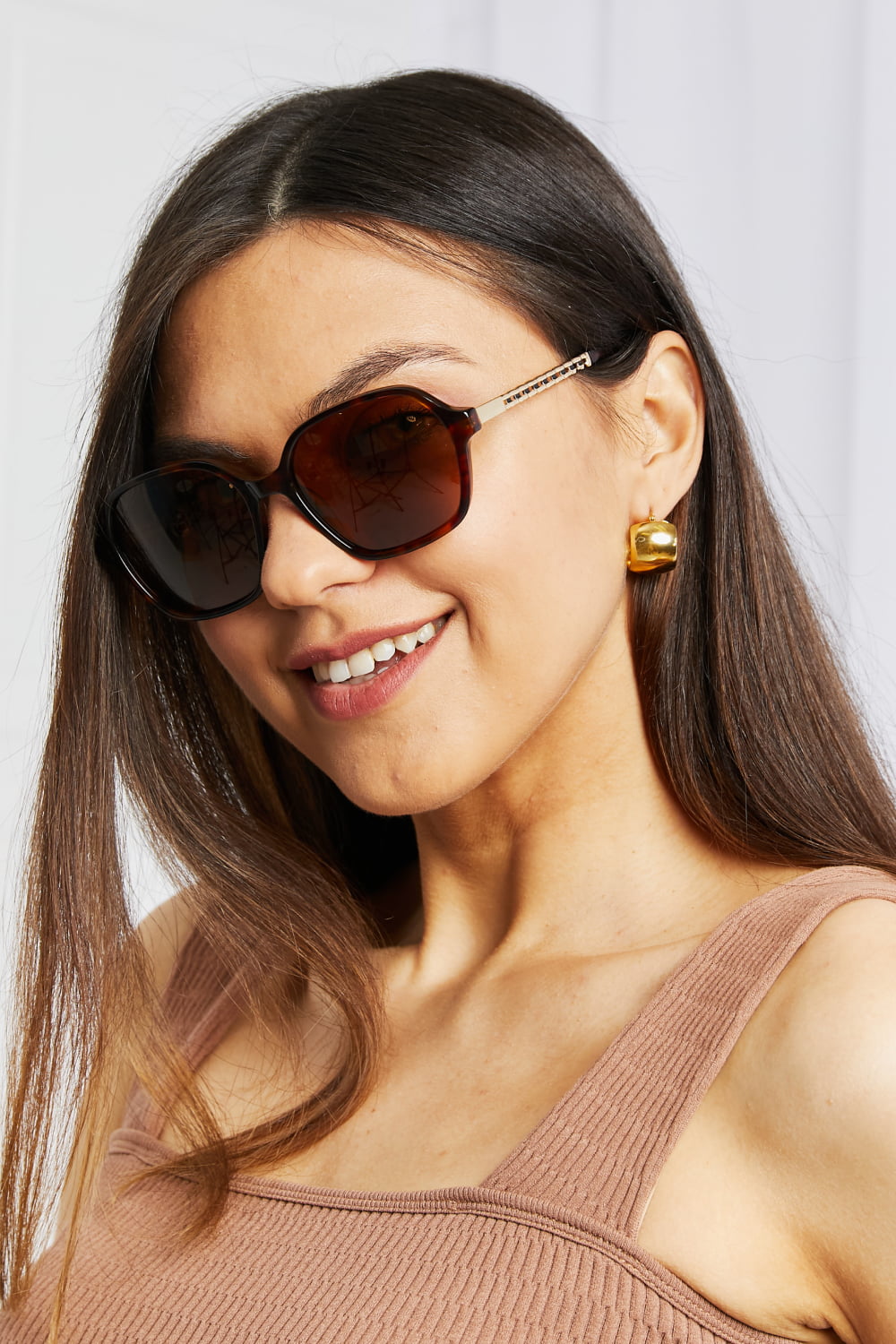 Smiling model tilts her head in brown full-rim, softly rounded square sunglasses with warm lenses and metal-acetate temples, wearing a ribbed sleeveless top.