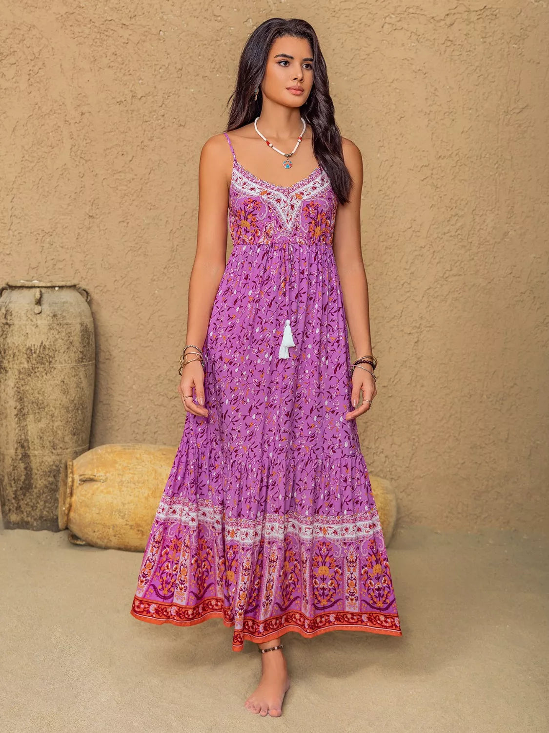 A model stands in a heliotrope purple floral print cami dress with delicate straps, a ruched bodice, flowing tiered maxi silhouette, tassel accent, and bordered hem.