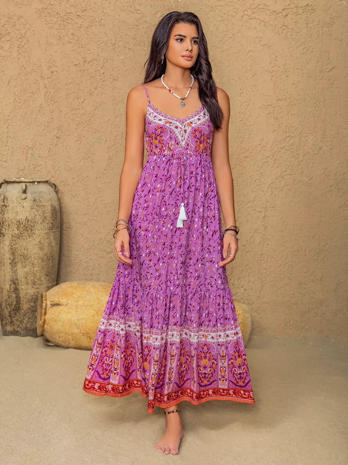 A model stands in a heliotrope purple floral print cami dress with delicate straps, a ruched bodice, flowing tiered maxi silhouette, tassel accent, and bordered hem.