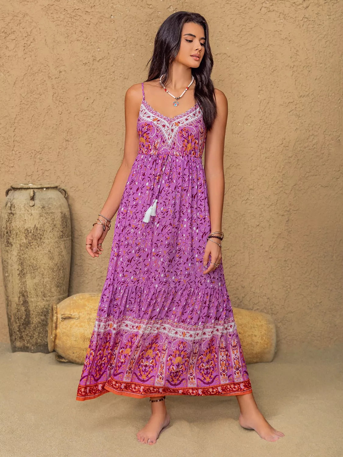 A model poses in a heliotrope purple floral print cami dress with delicate straps, a ruched bodice, tassel accents, flowing maxi silhouette, and bordered hem.