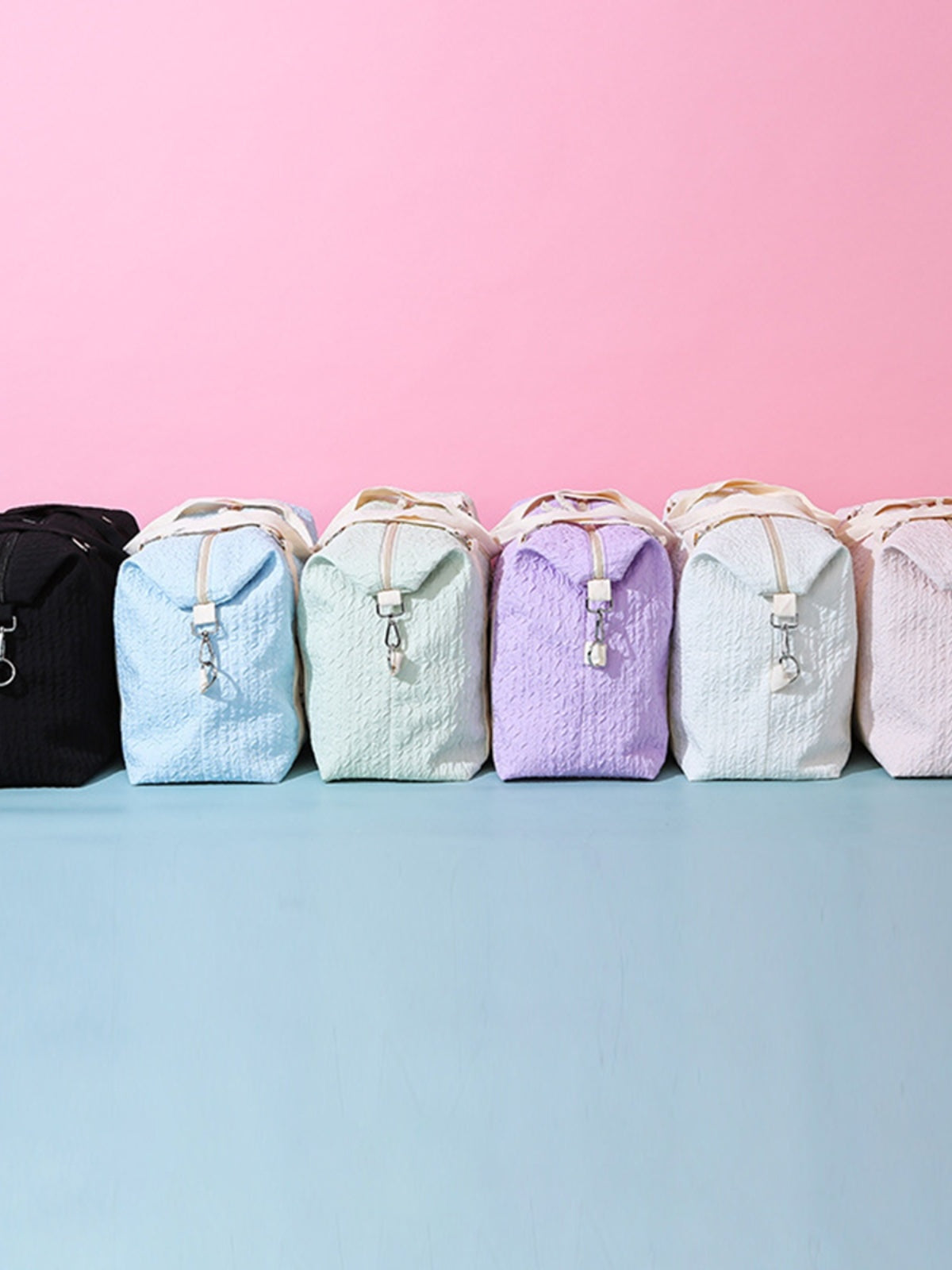 A row of textured Oxford cloth travel bags in black, blue, mint, lavender, white, and pink features softly structured silhouettes, top handles, and front strap details.