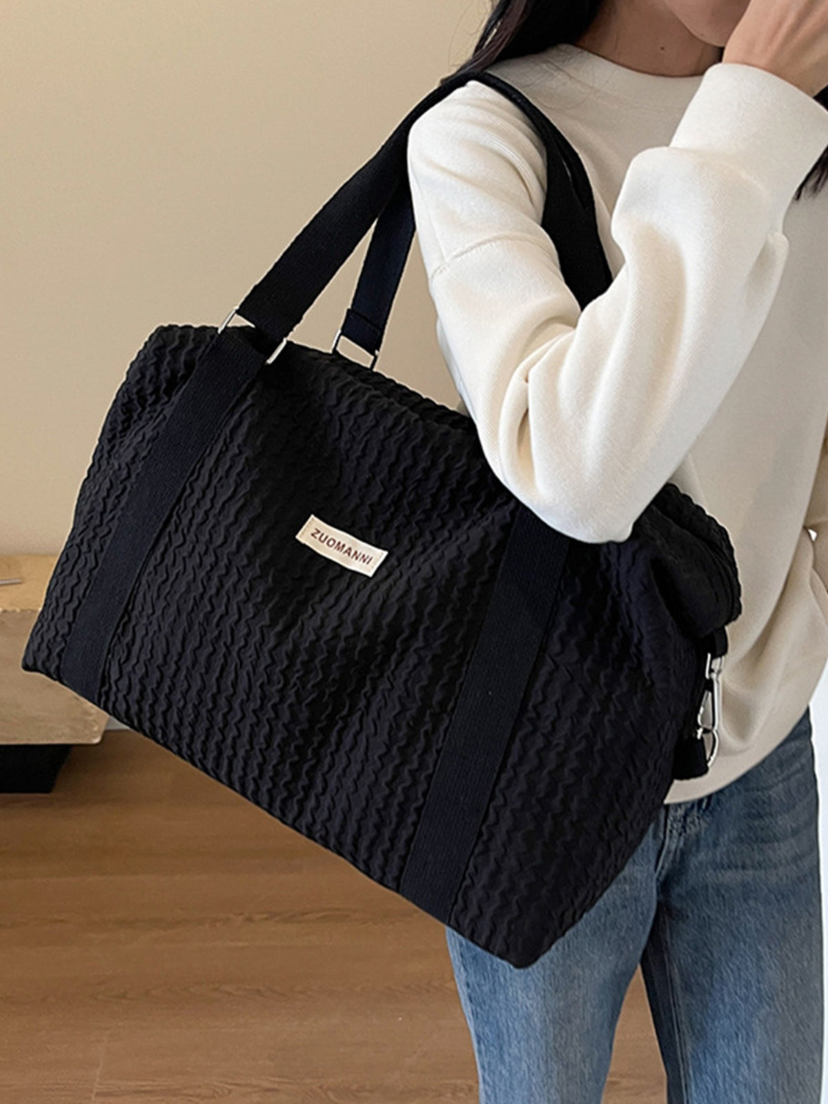 A model carries a large black textured Oxford cloth travel bag over one shoulder, featuring a softly structured silhouette, wide handles, and a detachable strap.