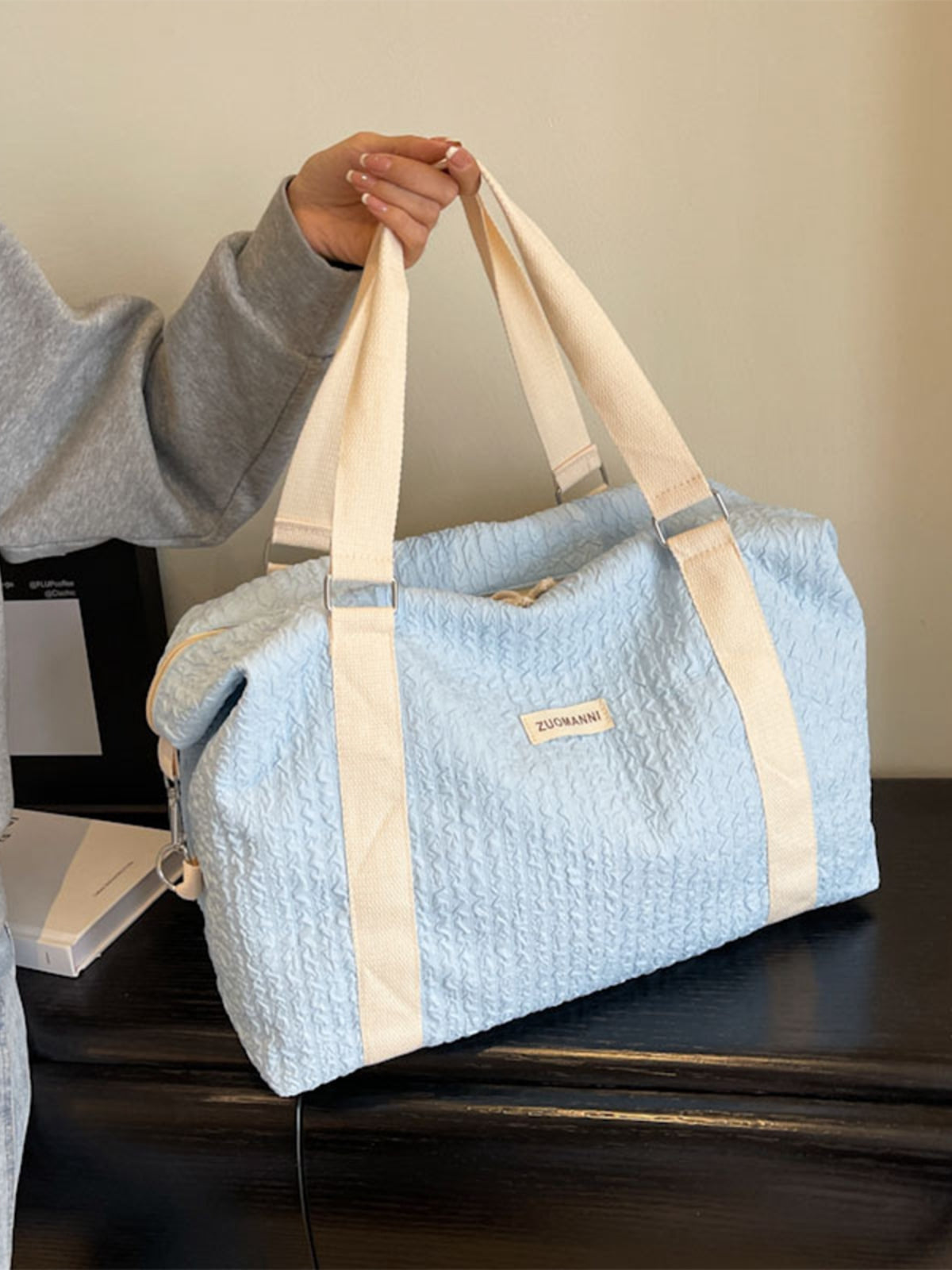 A model holds a large light blue textured Oxford cloth travel bag with a softly structured duffel silhouette, cream webbing handles, and silver hardware.