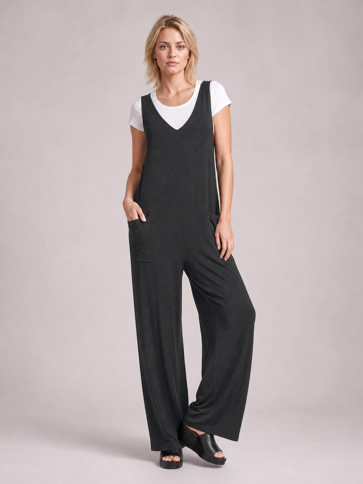 A model poses in a black textured sleeveless jumpsuit with a V-neck, wide-leg silhouette, front pockets, and a white T-shirt layered underneath.