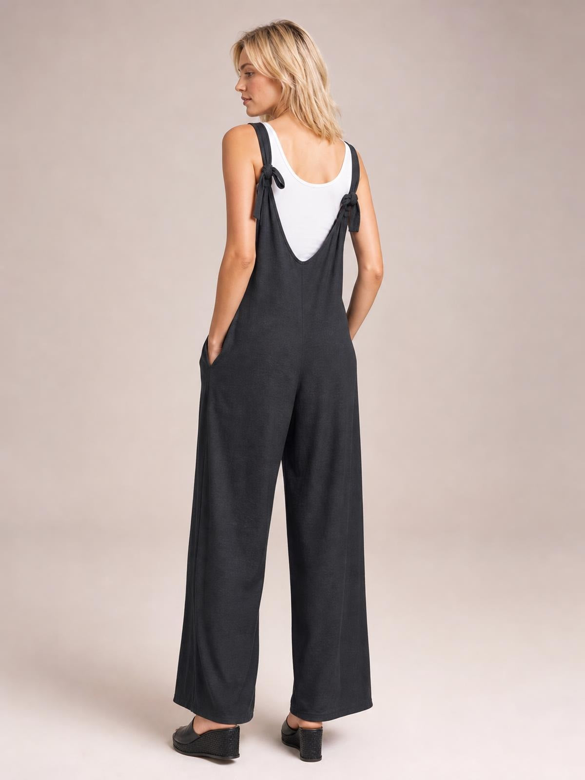 A model poses from the back in a charcoal textured sleeveless jumpsuit with tied shoulder straps, a deep V-back, roomy wide legs, and side pockets.