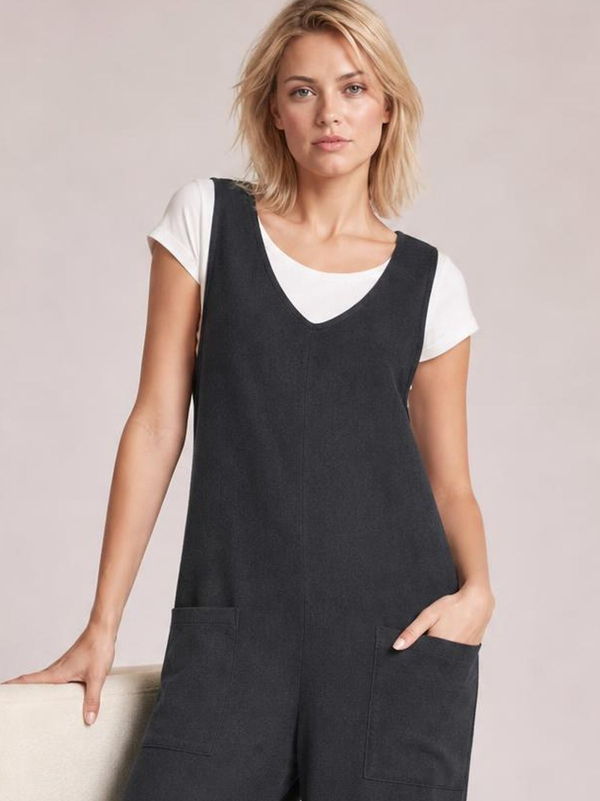 Model poses in a charcoal texture sleeveless jumpsuit with a V-neck, relaxed wide-leg silhouette, and large front patch pockets over a white tee.