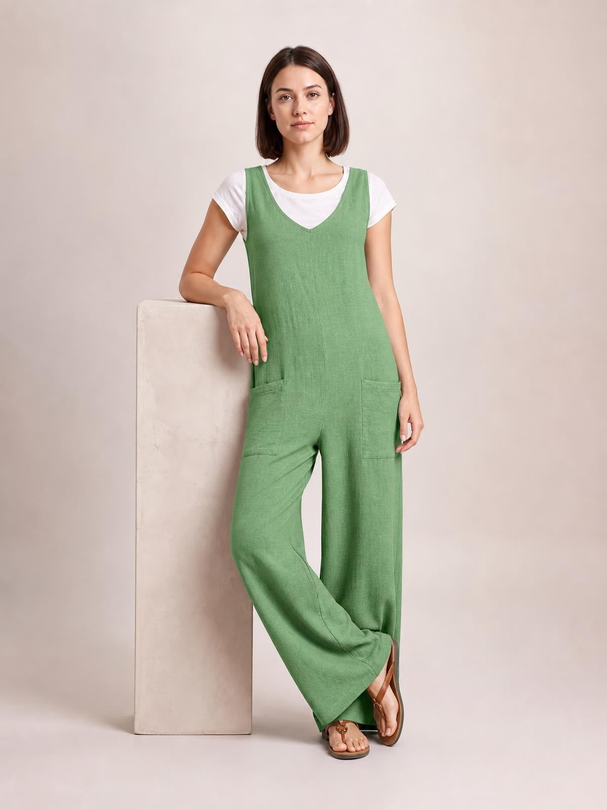 Model leans against a block wearing a sage green textured sleeveless jumpsuit with a V-neck, wide-leg silhouette, and two front pockets.