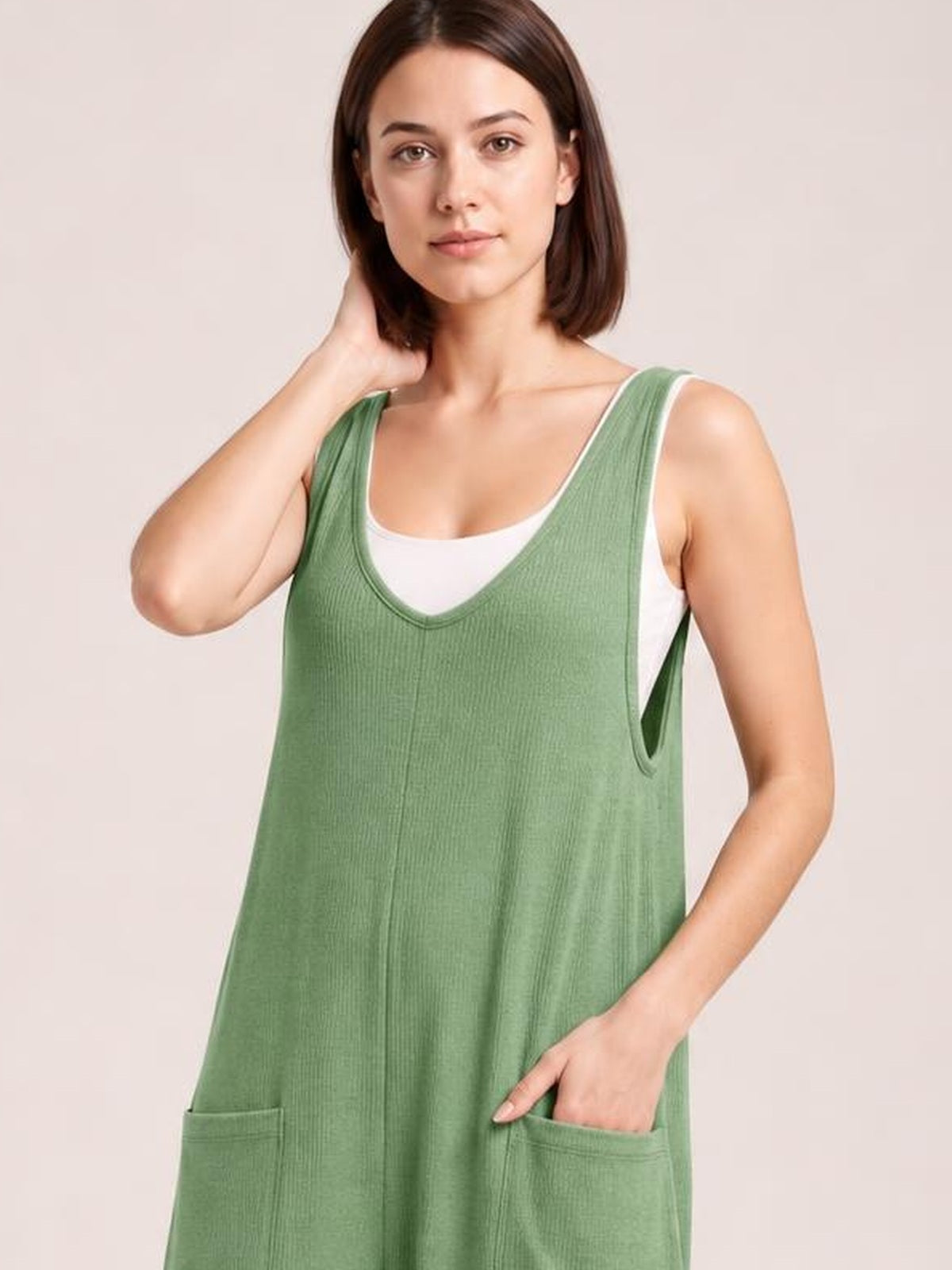 A model poses in a sage green textured sleeveless jumpsuit with a scoop neckline, relaxed silhouette, wide armholes, and front patch pockets.